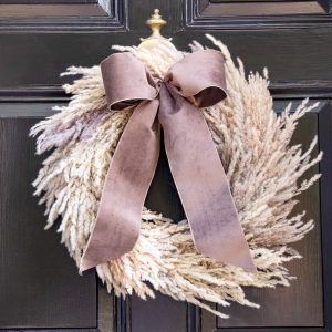 Pampas Grass Wreath for fall and winter made entirely of pampas grass plumes in it's natural color. It also features a dark taupe velvet ribbon with a deep brown taffeta lining. the wreath hangs on a front door that id black wood with a brass door knocker